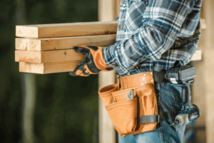 A man stands holding a wooden plank in his right hand, looking focused and ready to work on a project.