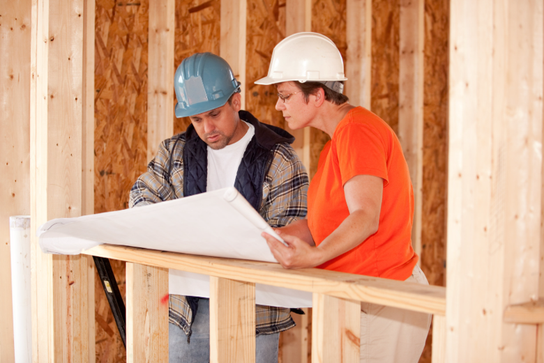 Two construction workers in blue and white hard hats review white blueprints inside a wood-framed room with tan wooden panels and exposed studs.